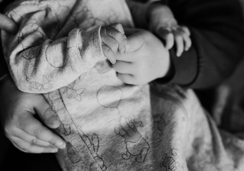 a black and white photo of a newborn baby and a toddler holding hands.